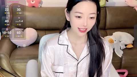 Media: A video of an East Asian woman with long black hair, wearing a white satin pajama set, sitting on a brown leather couch with plush toys, including a pink bear and a white duck.