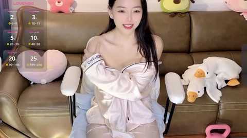 Media: Video of an Asian woman with long black hair, wearing a white satin off-shoulder robe, sitting on a beige leather couch. Background includes stuffed animals and a calendar overlay.