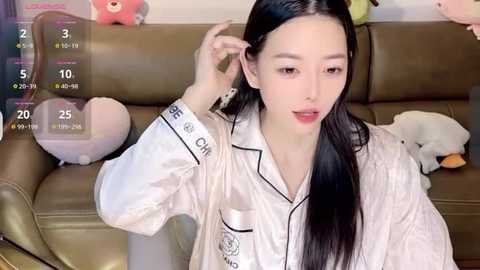 Media: Video of an East Asian woman with long black hair, light skin, and a slender figure, wearing a white satin robe with black trim, sitting on a beige leather couch. Background includes plush toys and a virtual date screen.