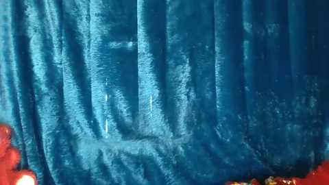 Media: Video of a blue velvet curtain with a textured surface, displaying slight creases and folds. The background is dimly lit, revealing faint shadows and a few reddish-brown objects in the lower part.