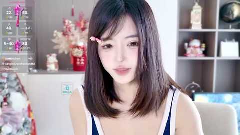 Media: Video of a young Asian woman with straight black hair, wearing a blue and white top, standing indoors. Background includes a wooden shelf with decorative items, and a digital screen displaying temperature and time.