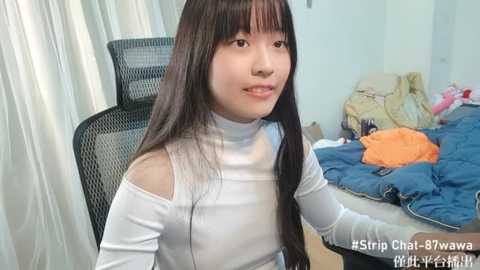 Media: Video of a young Asian woman with long black hair, wearing a white off-shoulder top, sitting on a black mesh chair in a messy bedroom with a blue bedspread.