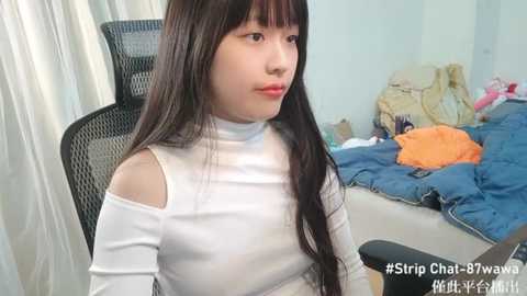 Media: Video of an Asian woman with long black hair in a white off-the-shoulder top, sitting in a black mesh chair. Background shows a messy bed with blue bedding and stuffed animals. Text overlay: \"#StripChat #Chaturbate #Asiandoll #AsiandollTV\".