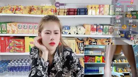 Media: Video of a young woman with light skin and blonde hair, wearing a black floral kimono, sitting in a brightly lit convenience store aisle filled with colorful snacks and beverages.