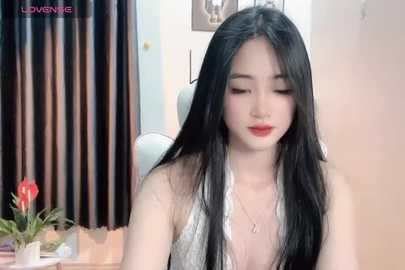 Media: Video of a young Asian woman with long black hair, fair skin, and red lipstick, wearing a silver halterneck top, sitting indoors with a potted red flower.