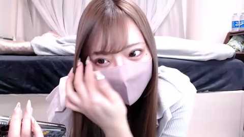 Video of an East Asian woman with long brown hair, wearing a white face mask, touching her nose, in a bedroom with a bed and nightstand in the background.