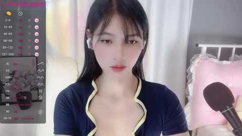 Media: Video of an East Asian woman with long black hair, wearing a navy blue cardigan, speaking into a microphone in a bedroom with a white bed and pink pillows.