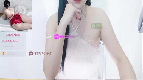 Media: Video of a slender, pale-skinned Asian woman with long black hair, wearing a white lace lingerie bra, lying on a bed, viewed from the waist up. Background includes a white wall and a \"LOUNIQUE\" advertisement.