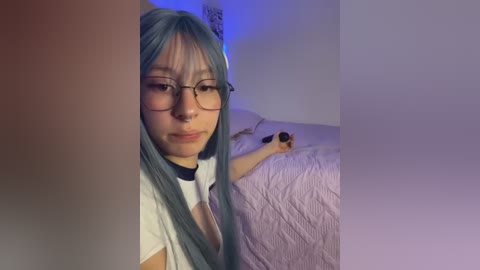 A young woman with long, pastel blue hair, wearing glasses, a black choker, and a white shirt, sits on a bed with a pink quilt. Her expression is neutral. The room is dimly lit with blue and purple lighting.