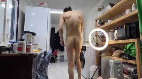 Media: Video of a nude man with fair skin, medium build, and short dark hair, standing in a cluttered, dimly lit bathroom. Surroundings include a sink, mirror, and open cabinet.