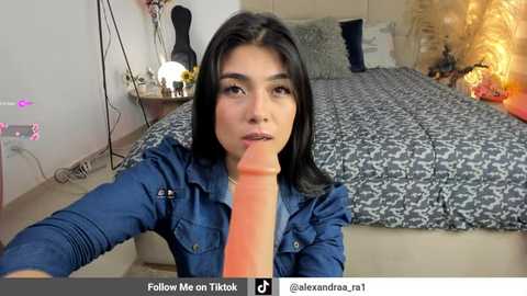 Video of a woman with long black hair, wearing a blue denim jacket, licking an erect, flesh-colored dildo. Background shows a bed with patterned bedding and a nightstand with a lamp.