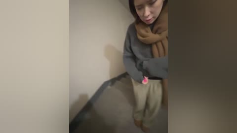 Media: A blurry video of a young person with medium skin tone, wearing a gray sweater, beige pants, and a brown scarf, standing in a beige hallway.