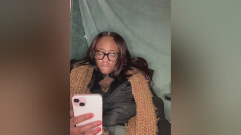 Media: Video of a Black woman with long, dark hair and glasses, wearing a black jacket, taking a selfie in a green tent.