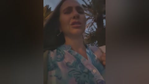 Media: Video of a woman with light skin, wearing a blue floral blouse, standing outdoors with blurred palm trees in the background.