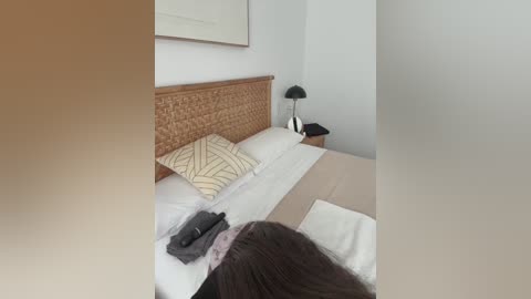 Video of a minimalist bedroom with a wicker headboard, white bedding, beige pillow with geometric pattern, black lamp on nightstand, person lying face down on bed, soft lighting.