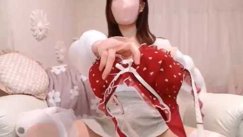 Media: Video of an Asian woman in a red polka-dotted bra, with long brown hair, sitting on a bed with white bedding and floral pillows in a pastel-toned room.