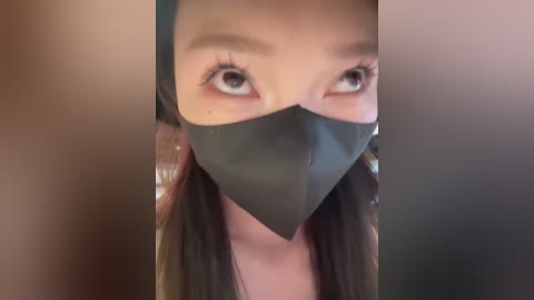 Media: Video of an Asian woman with long, dark hair, wearing a black face mask, showing only her eyes and nose. Background is blurred, with a beige and brown gradient.