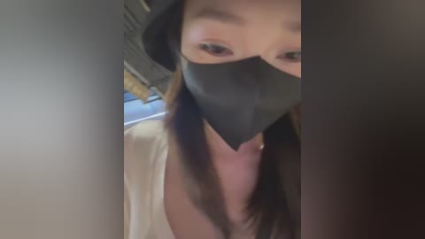 Media: Video of a person wearing a black surgical mask, with long brown hair, light skin, and a white top, taken indoors with a blurred background.