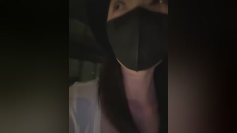 Media: A dimly lit video of a person wearing a black surgical mask, partially visible, with dark hair, likely indoors.