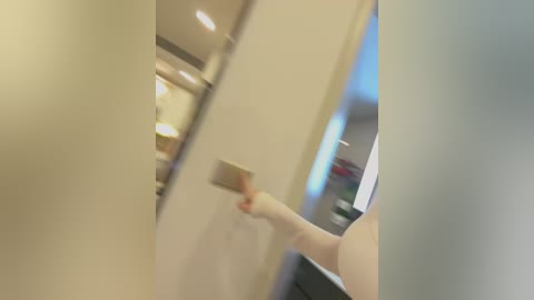 Media: A video shows a person, possibly female, with light skin, pressing a button on a sleek, beige wall in a modern, minimalist hallway. The background features a blurred, bright, open space with soft lighting.