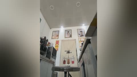 Media: Video of a small, well-lit kitchenette with white walls, stainless steel appliances, and a white stool. Three framed posters featuring coffee drinks adorn the wall, with a coffee maker and a milk frother on the counter.