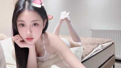 Media: Video of an Asian woman with long black hair, wearing a white lace bra and white gloves, lying on a bed in a minimalist room.