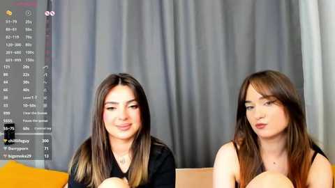 Media: Video of two young women with long brown hair, fair skin, and light makeup, wearing black tops, seated side by side, in front of a grey curtain background.