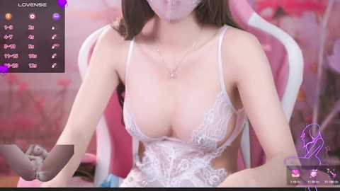 Media: Video of a light-skinned, slender Asian woman with medium-sized breasts wearing a sheer, lace-trimmed white dress, sitting on a pink gaming chair in a room with pink floral wallpaper.