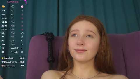 Media: A video of a young Caucasian woman with long, straight red hair, fair skin, and a slight smile, sitting in a purple chair. The background features teal curtains.