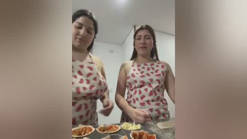 Media: Video of two women in a kitchen, one with large breasts, wearing a red and white polka dot apron, mixing ingredients on a granite countertop.