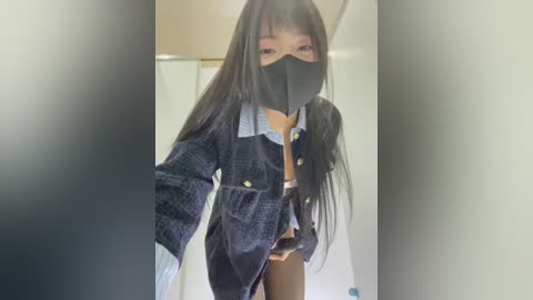 Video of a slender Asian woman with long black hair, wearing a black face mask, blue denim jacket, and black leggings, standing in a bathroom with a blurred background.