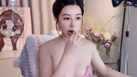 Media: Video of an East Asian woman with fair skin, dark hair, and small breasts, wearing a pink bra, licking her finger, in a cozy, floral-decorated room.