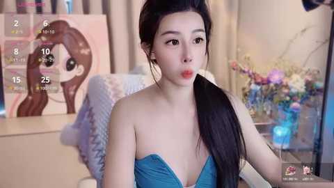 Media: A video of an Asian woman with long black hair, wearing a strapless blue dress, sitting indoors, holding a phone. The background features a calendar, a flower arrangement, and a pastel-colored wall.