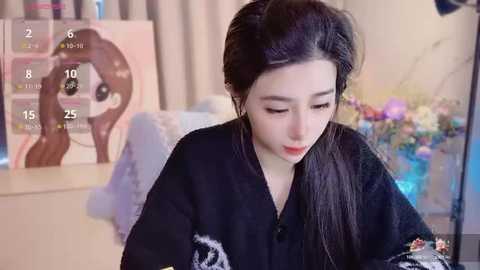 Media: A video of an East Asian woman with long black hair in a black cardigan, sitting in a cozy room with a colorful floral painting and a soft-focus background.