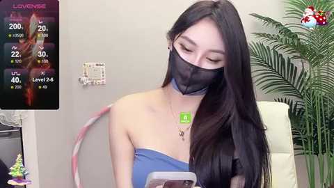 Video of an East Asian woman with long black hair, wearing a blue strapless dress, black face mask, and a green name tag, seated indoors with a potted plant and a digital screen displaying a fitness app.