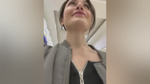 Video of a young woman with fair skin, brown hair, and light makeup, wearing a gray jacket and black top, smiling and looking up. Background shows a ceiling and part of a wall.