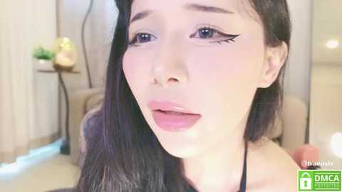 Media: Video of a young Asian woman with fair skin, long black hair, and sharp winged eyeliner, wearing a black top, in a modern living room with beige walls, a potted plant, and a dimly lit background.