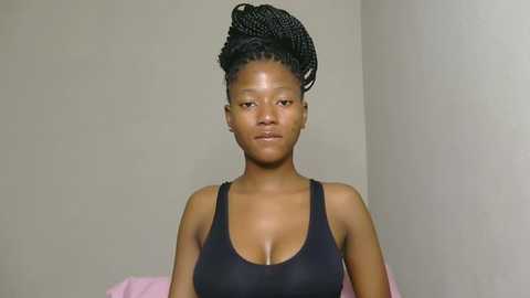 Video of a Black woman with medium-dark skin and a slender build, wearing a black sports bra. She has her hair styled in long, twisted braids. The background is plain and beige.