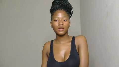 Media: Video of a dark-skinned woman with a tall, elaborate braided hairstyle, wearing a black tank top, standing against a plain, beige wall.