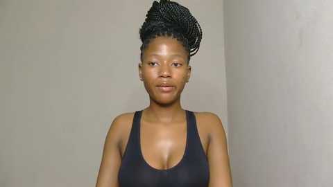 Video of a dark-skinned African woman with a medium build, wearing a black tank top and styled hair in a high, intricate braid. She stands against a plain beige wall, appearing solemn and composed.