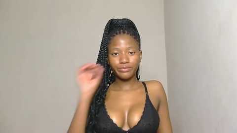 Video of a dark-skinned woman with braided hair, wearing a black lace bra, posing against a plain beige wall. She is smiling and lifting her right hand.