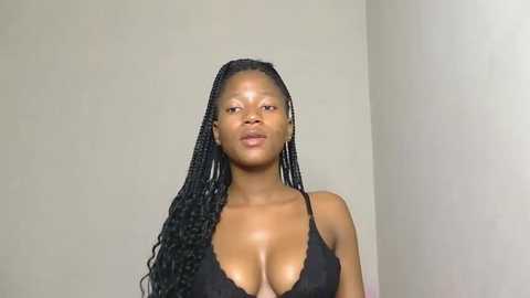 Media: Video of a slender, dark-skinned woman with long, braided hair, wearing a black lace bra. She stands against a plain, light-colored wall, exuding a confident expression.