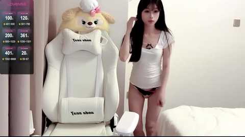 Video of a slim East Asian woman with long black hair, wearing a white tank top and black panties, standing next to a white gaming chair.