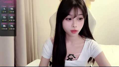 Media: Video of a young East Asian woman with long black hair, wearing a white shirt and pink bra, sitting on a white chair in a dimly lit room with beige curtains.