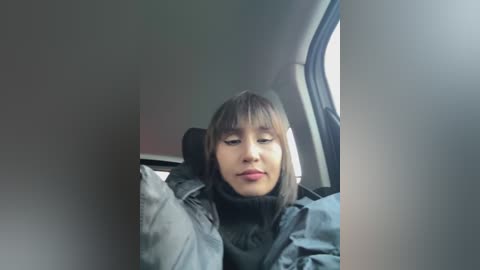 Media: Video of a young woman with light skin, straight black hair, and bangs, wearing a black coat and scarf, sitting in a car with a window view of a cityscape.