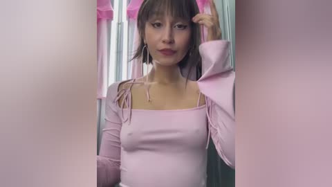 Media: Video of a young Asian woman with short, dark hair and pale skin, wearing a light pink, long-sleeve top and pink headband. She stands in front of a pink, blurry background.