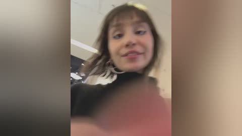 Media: A blurry video of a young woman with light brown hair and a neutral expression, wearing a black top, taken indoors.