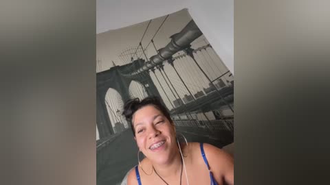 Media: Video of a smiling Latina woman with dark hair in a bun, wearing a blue bra, in front of a large mural of the Brooklyn Bridge.