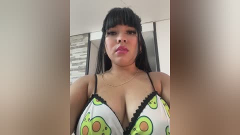 Video of a young woman with long black hair and bangs, wearing a white crop top adorned with avocado print and black trim. She has a curvy physique, medium skin tone, and full lips. Background shows a modern room with a textured wall.