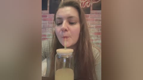 Media: Video of a light-skinned woman with long brown hair, wearing a beige sweater, drinking from a mason jar with a wooden lid, against a red brick wall background.